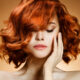 Close-up portrait of a woman with vivid red curly hair, eyes closed, hand resting on her cheek against a warm orange background.