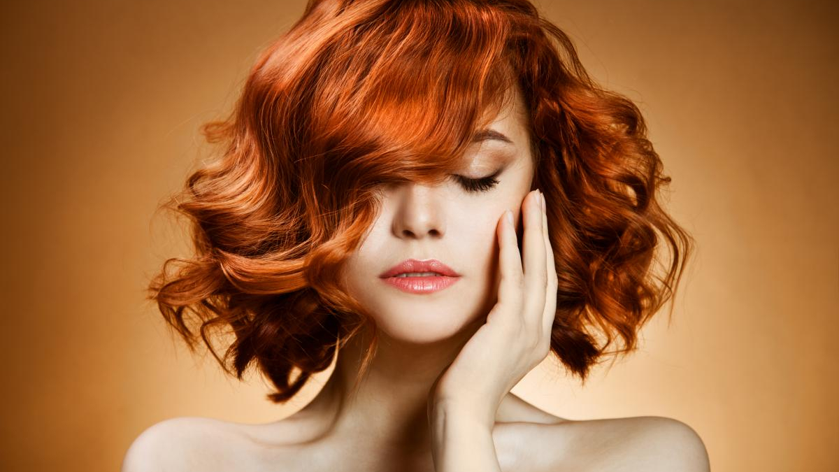 Close-up portrait of a woman with vivid red curly hair, eyes closed, hand resting on her cheek against a warm orange background.