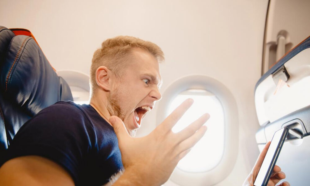 Man on an airplane screams in alarm while looking at a smartphone held in his hand on the tray table side.
