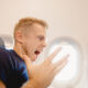 Man on an airplane screams in alarm while looking at a smartphone held in his hand on the tray table side.