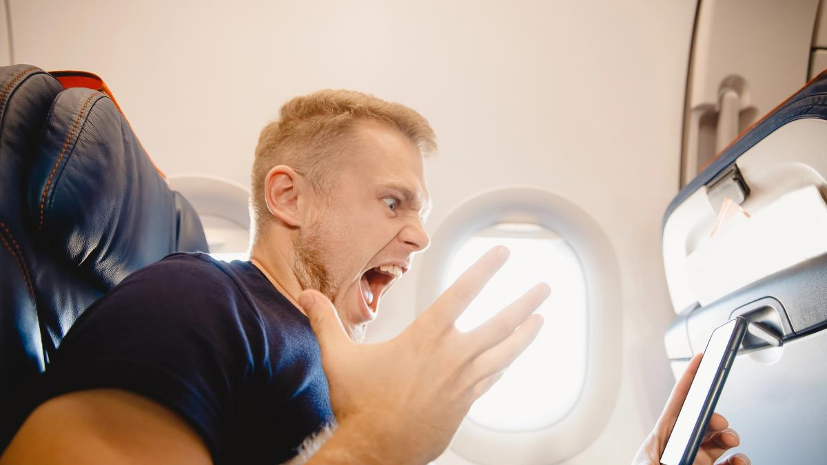 Man on an airplane screams in alarm while looking at a smartphone held in his hand on the tray table side.