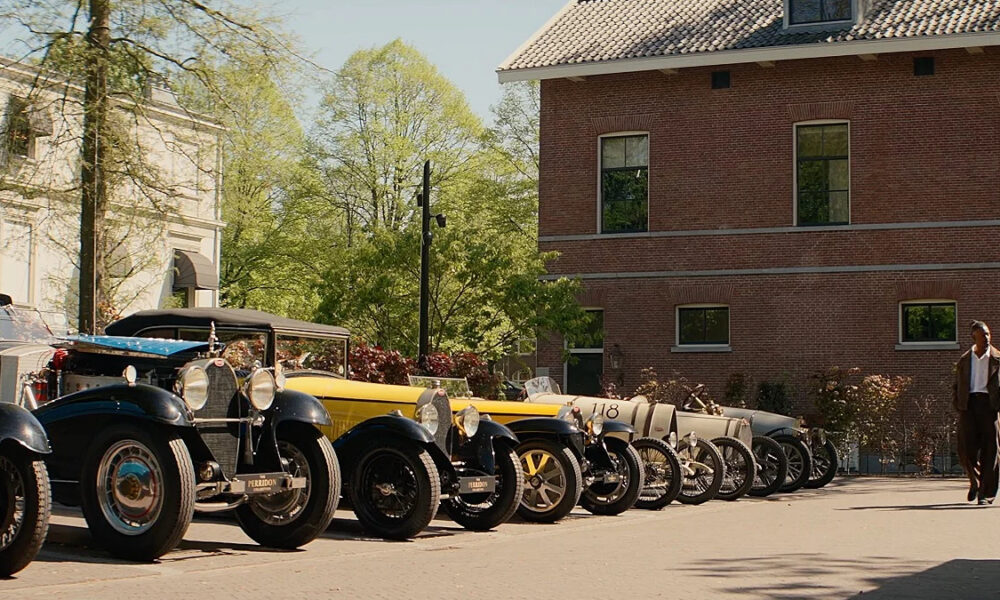 Row of vintage cars parked along a sunny street beside a brick building; two men walk toward the camera in the distance.