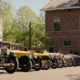 Row of vintage cars parked along a sunny street beside a brick building; two men walk toward the camera in the distance.