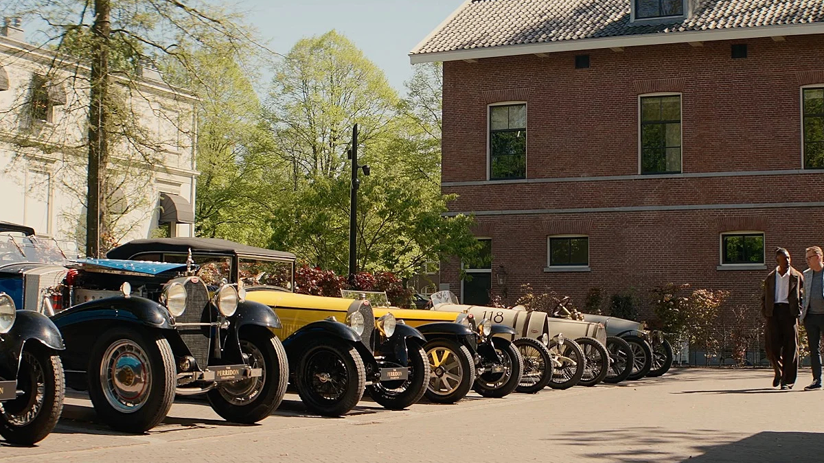 Row of vintage cars parked along a sunny street beside a brick building; two men walk toward the camera in the distance.