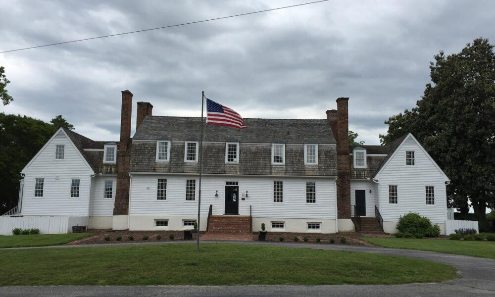 White two-story colonial house with dormer windows, brick chimneys, and a central door, flanked by steps and a flagpole with an American flag in front.