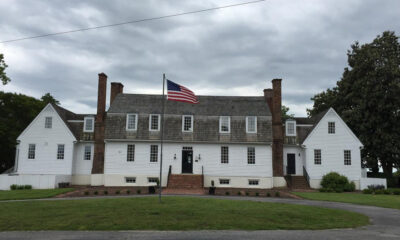 White two-story colonial house with dormer windows, brick chimneys, and a central door, flanked by steps and a flagpole with an American flag in front.