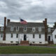 White two-story colonial house with dormer windows, brick chimneys, and a central door, flanked by steps and a flagpole with an American flag in front.