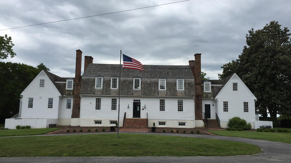 White two-story colonial house with dormer windows, brick chimneys, and a central door, flanked by steps and a flagpole with an American flag in front.