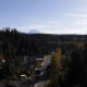 Aerial view of a small town along a main road bordered by dense evergreen trees, with a distant snow-capped mountain under a clear blue sky.