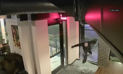 A person in a dark hoodie reaches toward the glass door at the U Wine Bar entrance, as if attempting to break in, at night outside.