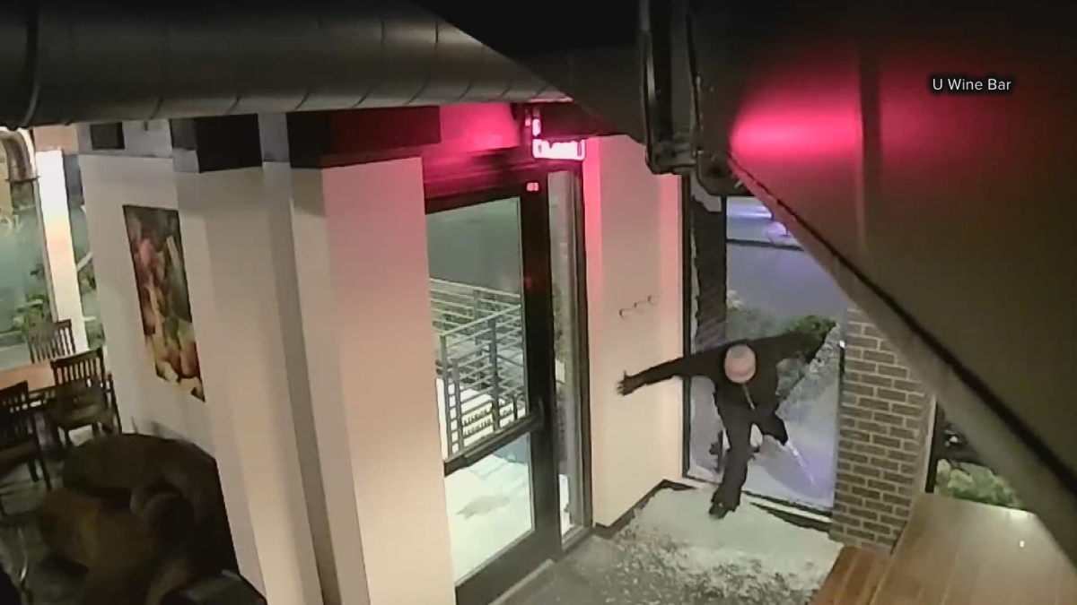 A person in a dark hoodie reaches toward the glass door at the U Wine Bar entrance, as if attempting to break in, at night outside.