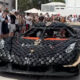 Exotic black sports car wrapped in circular panels on display, surrounded by a crowd of photographers and onlookers.