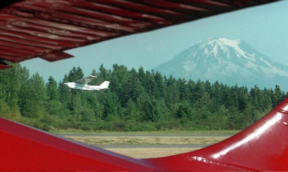 Small white airplane flying over a forest with a snow-capped mountain in the distance, viewed between red aircraft wings in the foreground.