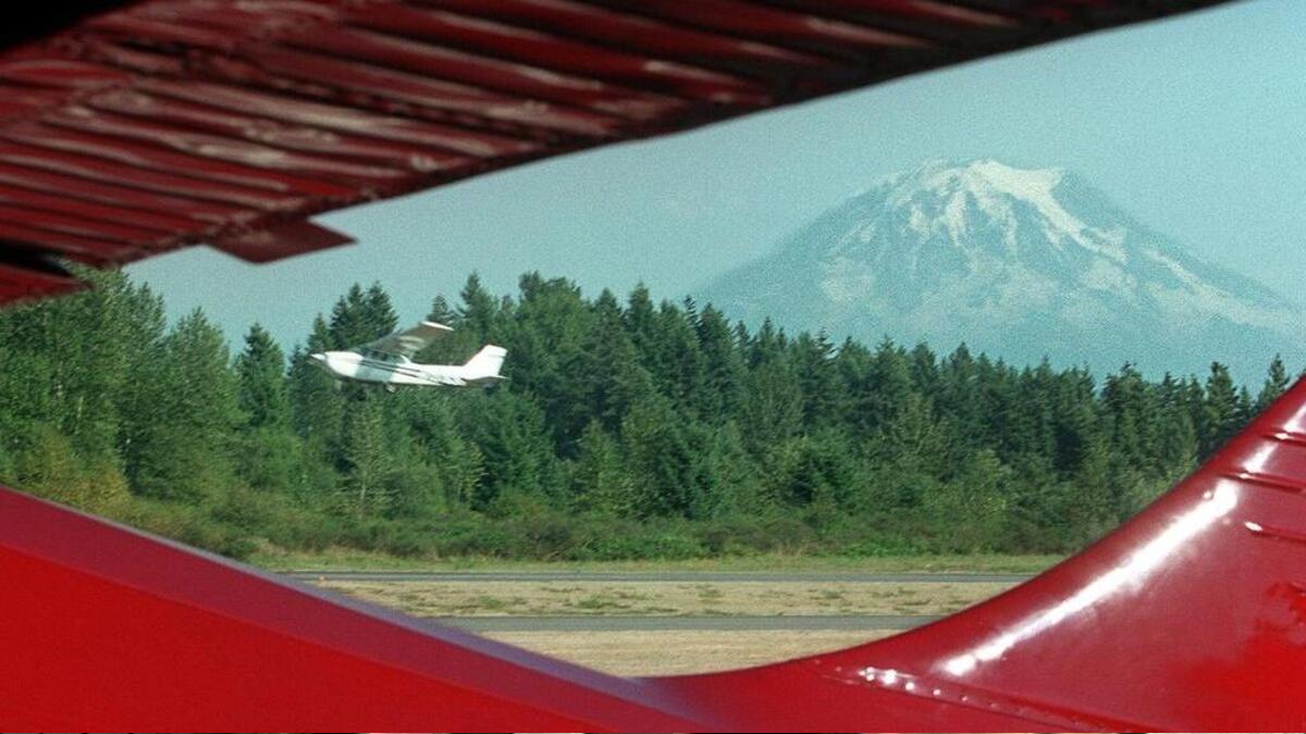 Small white airplane flying over a forest with a snow-capped mountain in the distance, viewed between red aircraft wings in the foreground.