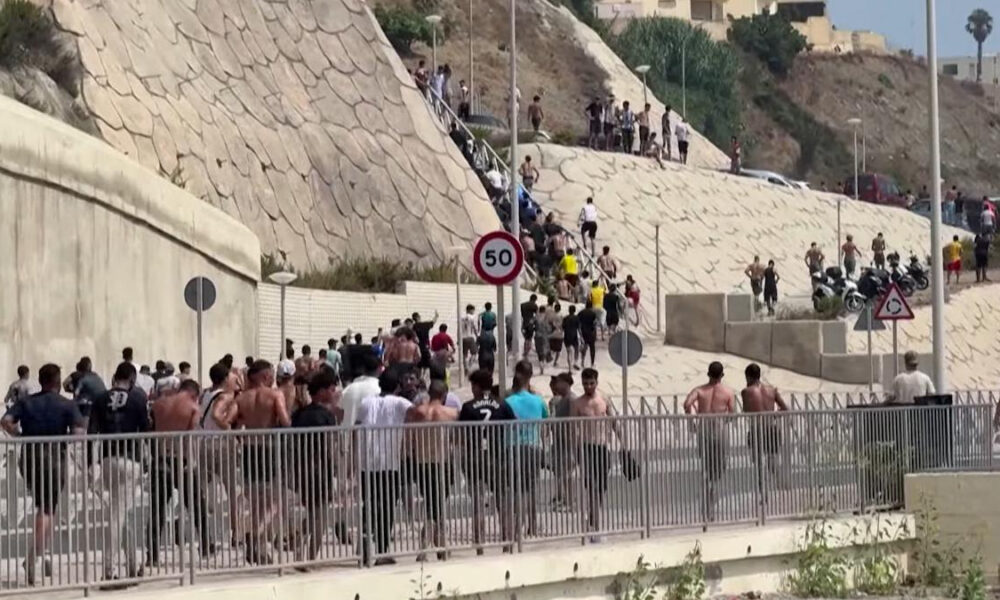 Large group of people running along a barricaded street beside a stone hillside, many shirtless, with a 50 km/h sign in view.
