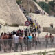 Large group of people running along a barricaded street beside a stone hillside, many shirtless, with a 50 km/h sign in view.