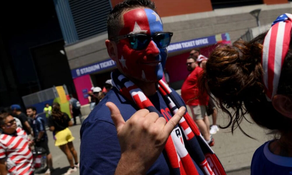 Man with red, white, and blue star face paint wearing sunglasses gives a thumbs-up at a sports event outside a stadium.