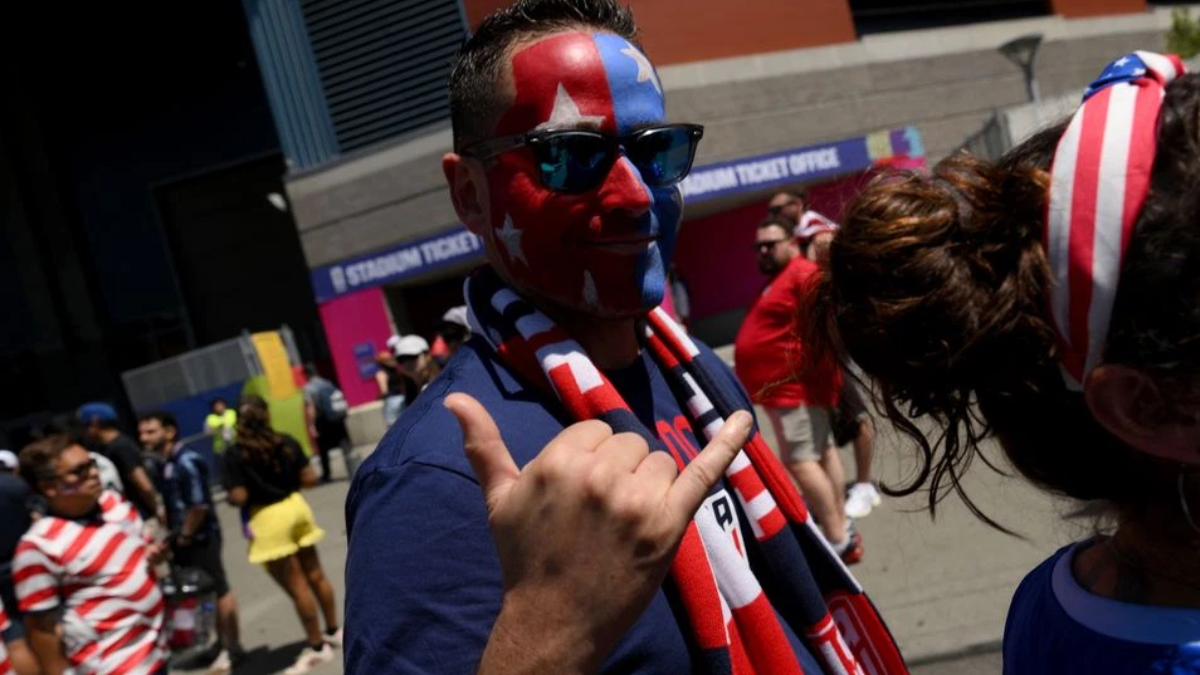 Man with red, white, and blue star face paint wearing sunglasses gives a thumbs-up at a sports event outside a stadium.