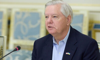 Middle-aged man in a navy blazer and light blue shirt speaks into a microphone, wearing a US-Ukraine flag pin on his lapel.