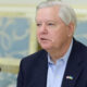 Middle-aged man in a navy blazer and light blue shirt speaks into a microphone, wearing a US-Ukraine flag pin on his lapel.
