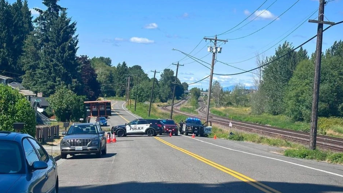 Police cars block a rural two-lane road with orange cones; railroad tracks and trees in the background.