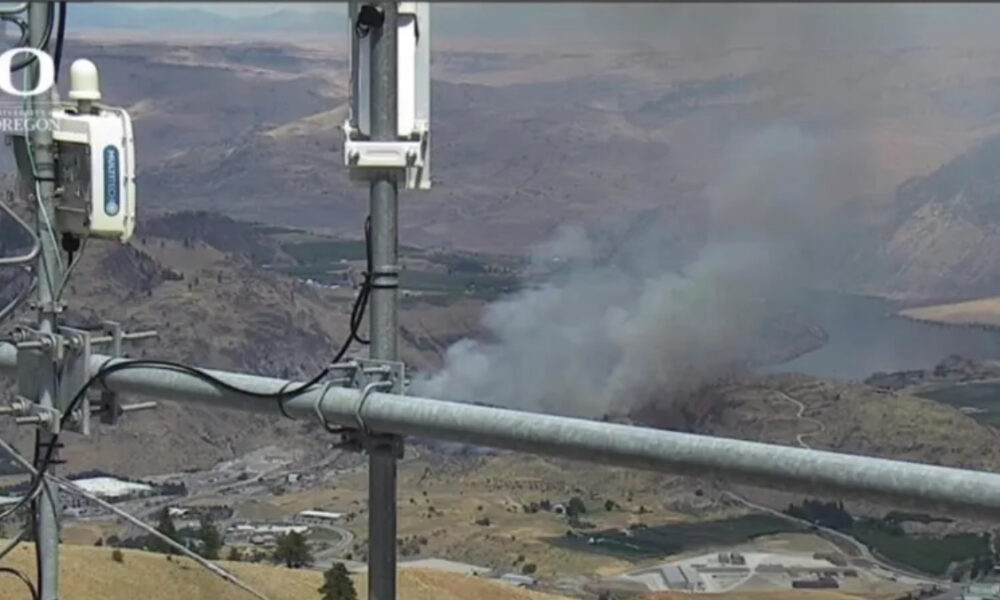 Electrical transmission equipment in the foreground with a smoke plume rising from a hillside fire in a valley below.