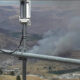 Electrical transmission equipment in the foreground with a smoke plume rising from a hillside fire in a valley below.