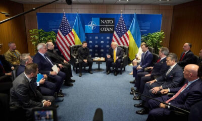 Group of officials in a U-shaped seating arrangement for a NATO meeting, with U.S. and Ukrainian flags behind.
