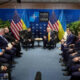 Group of officials in a U-shaped seating arrangement for a NATO meeting, with U.S. and Ukrainian flags behind.