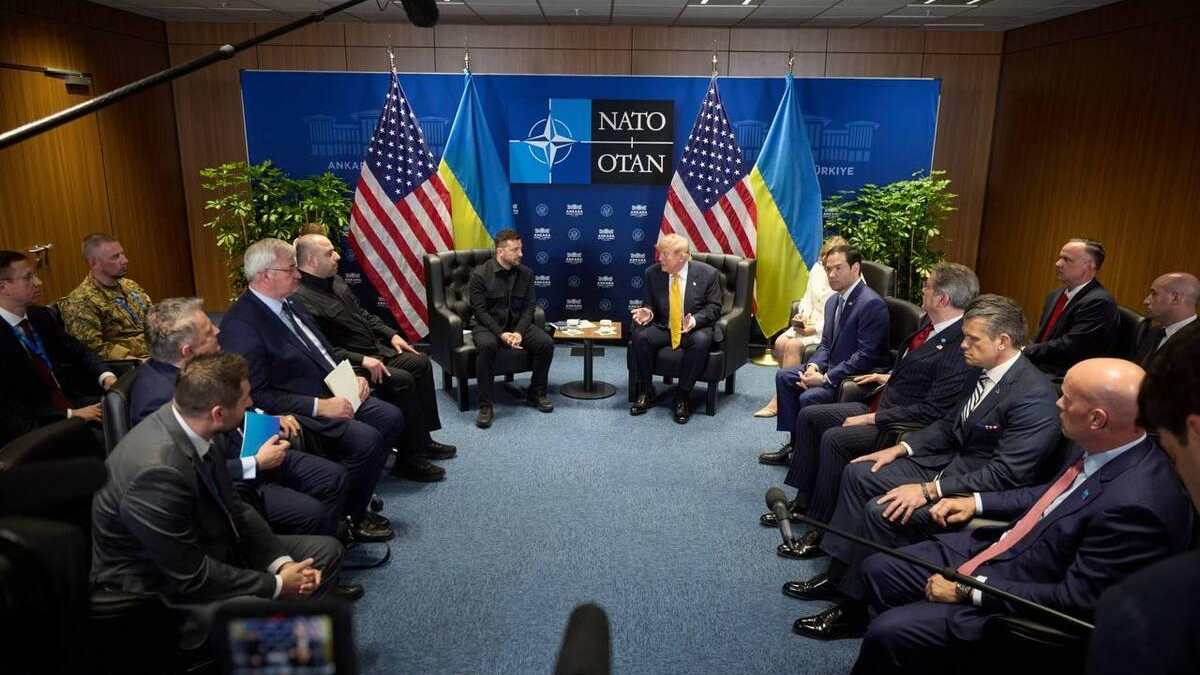 Group of officials in a U-shaped seating arrangement for a NATO meeting, with U.S. and Ukrainian flags behind.
