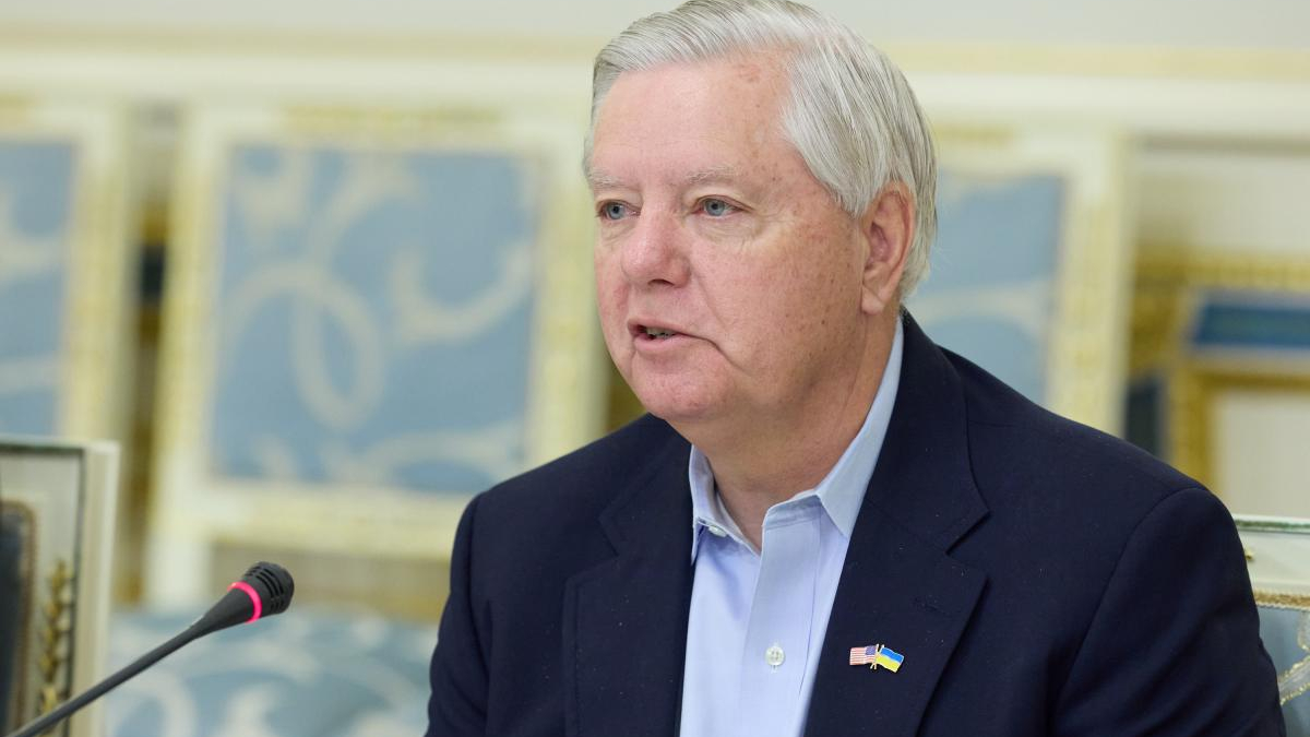 Mid-speech portrait of a man in a navy blazer and light blue shirt, wearing US and Ukraine pins, speaking at a microphone in a formal setting.