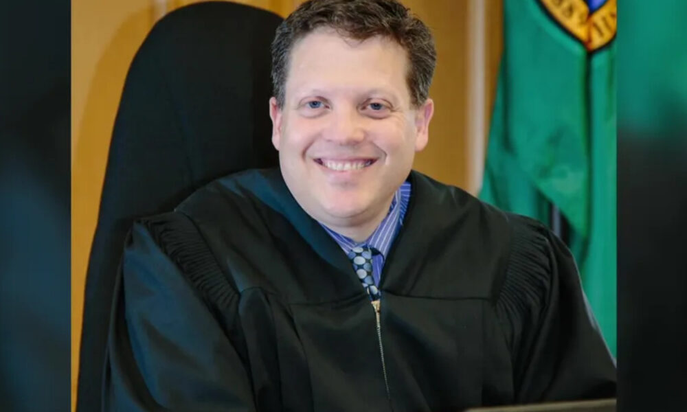 Smiling judge in a black robe seated in a courtroom with a flag in the background.