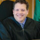 Smiling judge in a black robe seated in a courtroom with a flag in the background.
