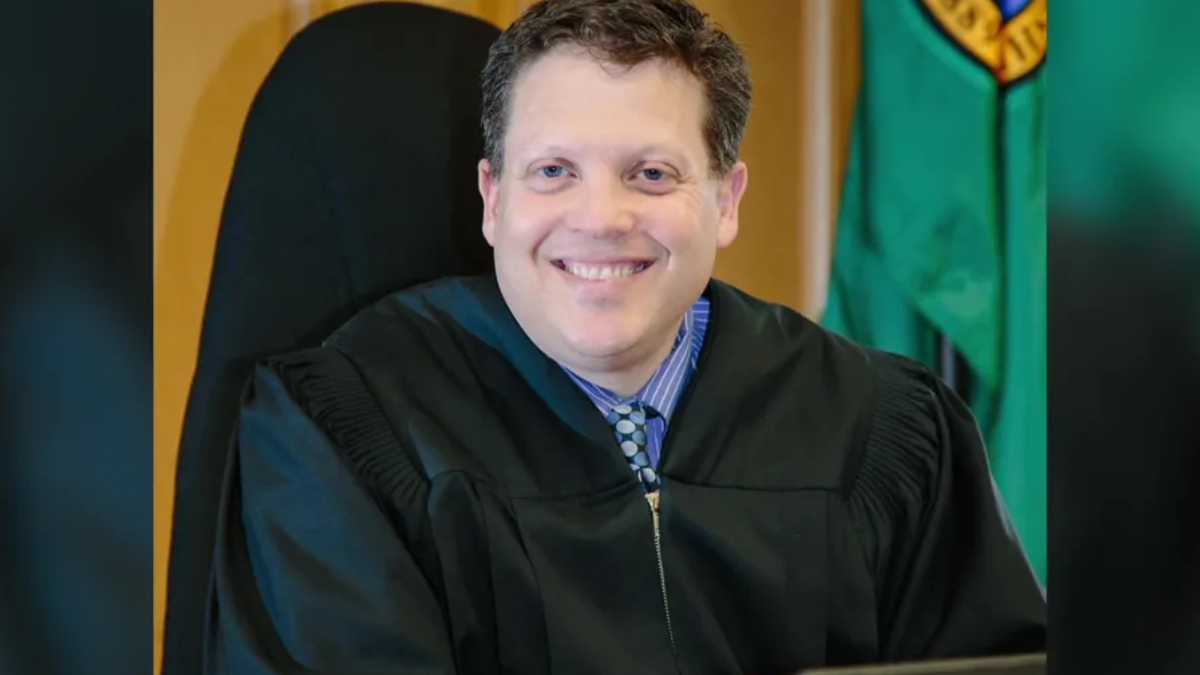 Smiling judge in a black robe seated in a courtroom with a flag in the background.