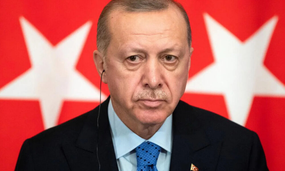 Close-up of a serious man in a dark suit and blue tie wearing an ear microphone, red background with white stars.