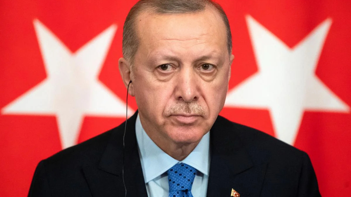 Close-up of a serious man in a dark suit and blue tie wearing an ear microphone, red background with white stars.
