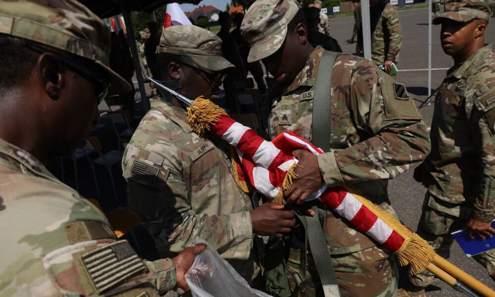 Soldiers in camouflage handle a folded American flag with gold tassels during a ceremony at an outdoor event.