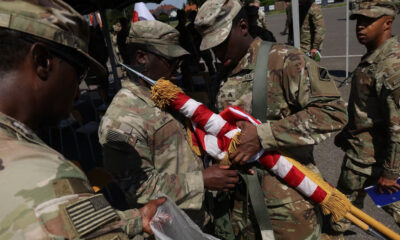 Soldiers in camouflage handle a folded American flag with gold tassels during a ceremony at an outdoor event.