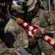 Soldiers in camouflage handle a folded American flag with gold tassels during a ceremony at an outdoor event.