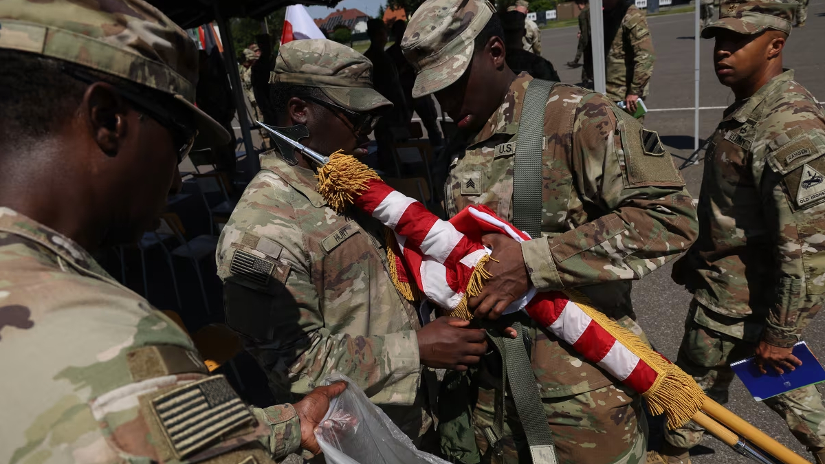 Soldiers in camouflage handle a folded American flag with gold tassels during a ceremony at an outdoor event.