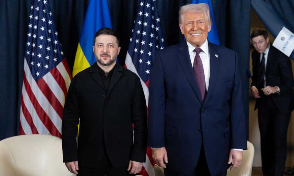 Ukrainian President Zelenskyy and former President Trump pose for a photo at a formal event with US and Ukrainian flags behind them.
