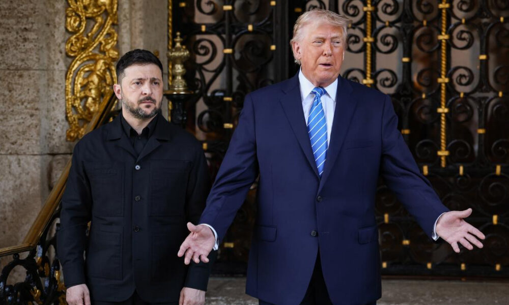 Man in a navy suit with a blue striped tie gestures with open hands beside a security guard outside ornate gates.