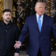 Man in a navy suit with a blue striped tie gestures with open hands beside a security guard outside ornate gates.