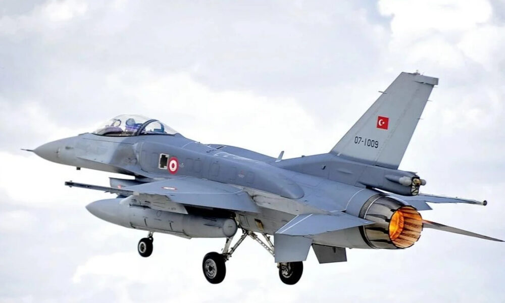 Turkish Air Force two-seat fighter jet in flight with afterburner flame, gray aircraft against a cloudy sky.