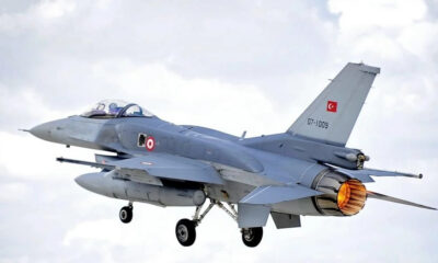 Turkish Air Force two-seat fighter jet in flight with afterburner flame, gray aircraft against a cloudy sky.