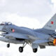Turkish Air Force two-seat fighter jet in flight with afterburner flame, gray aircraft against a cloudy sky.
