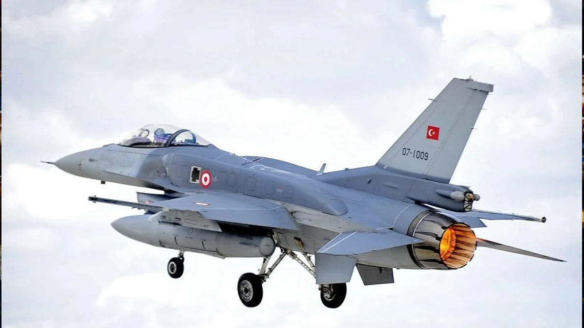 Turkish Air Force two-seat fighter jet in flight with afterburner flame, gray aircraft against a cloudy sky.