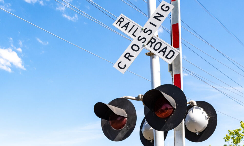 Railroad crossing sign reading 'RAILROAD CROSSING' with red warning lights on a metal pole against a blue sky and power lines behind it.