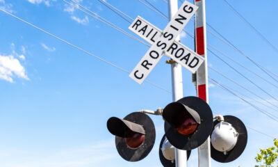 Railroad crossing sign reading 'RAILROAD CROSSING' with red warning lights on a metal pole against a blue sky and power lines behind it.