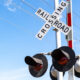 Railroad crossing sign reading 'RAILROAD CROSSING' with red warning lights on a metal pole against a blue sky and power lines behind it.